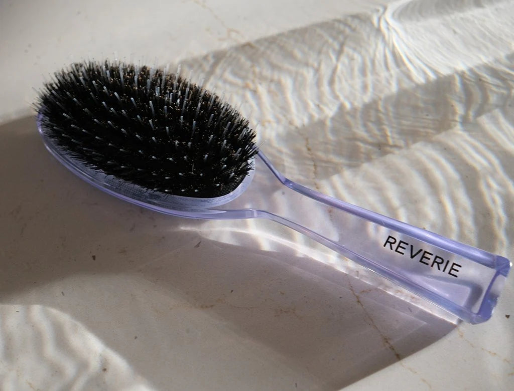 Hairbrush with black bristles and a purple handle labeled 'REVERIE' on a textured white surface.