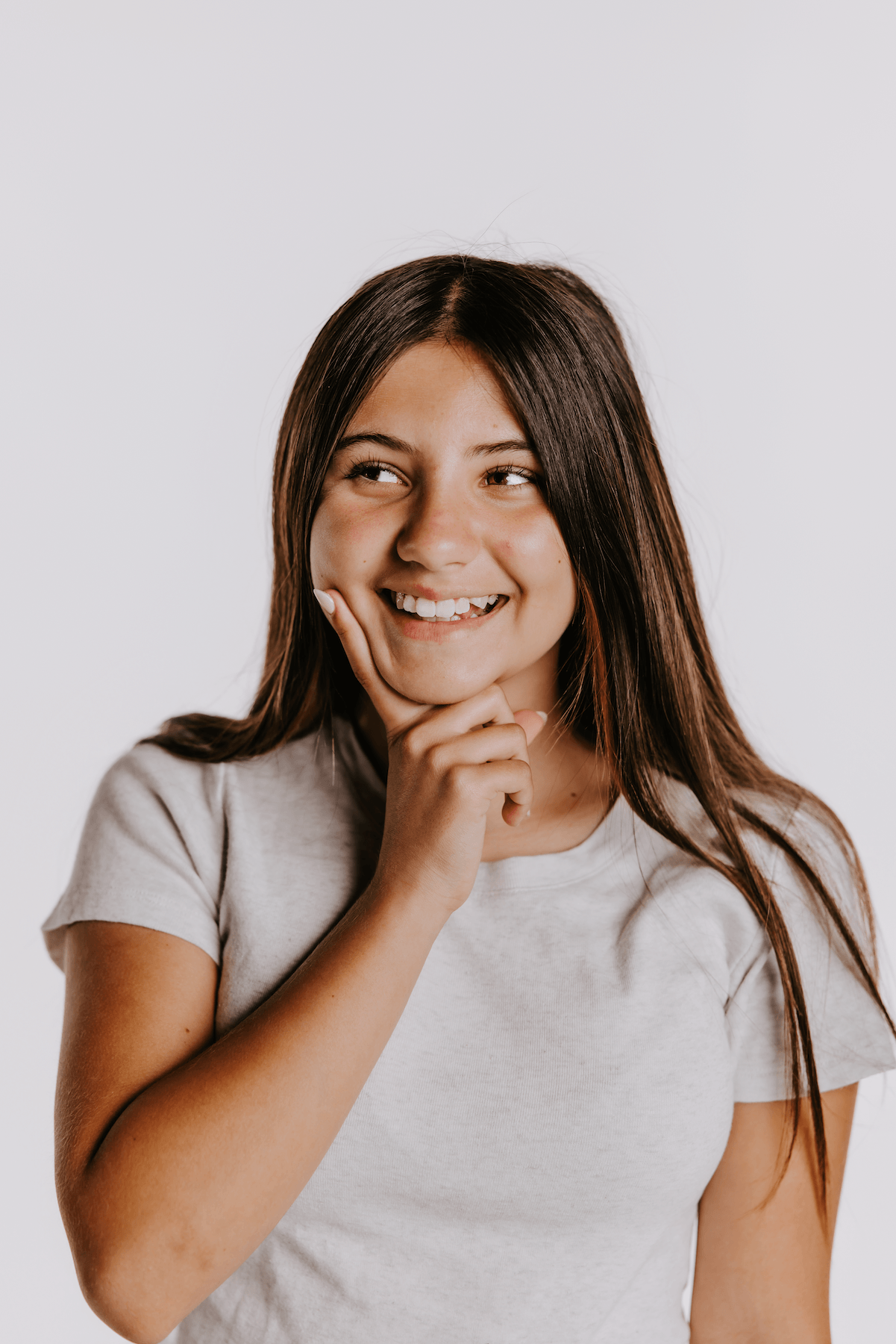 A smiling young girl with long dark brown hair looks to the side while resting her chin on her hand, wearing a light gray crew-neck t-shirt against a plain light gray background.