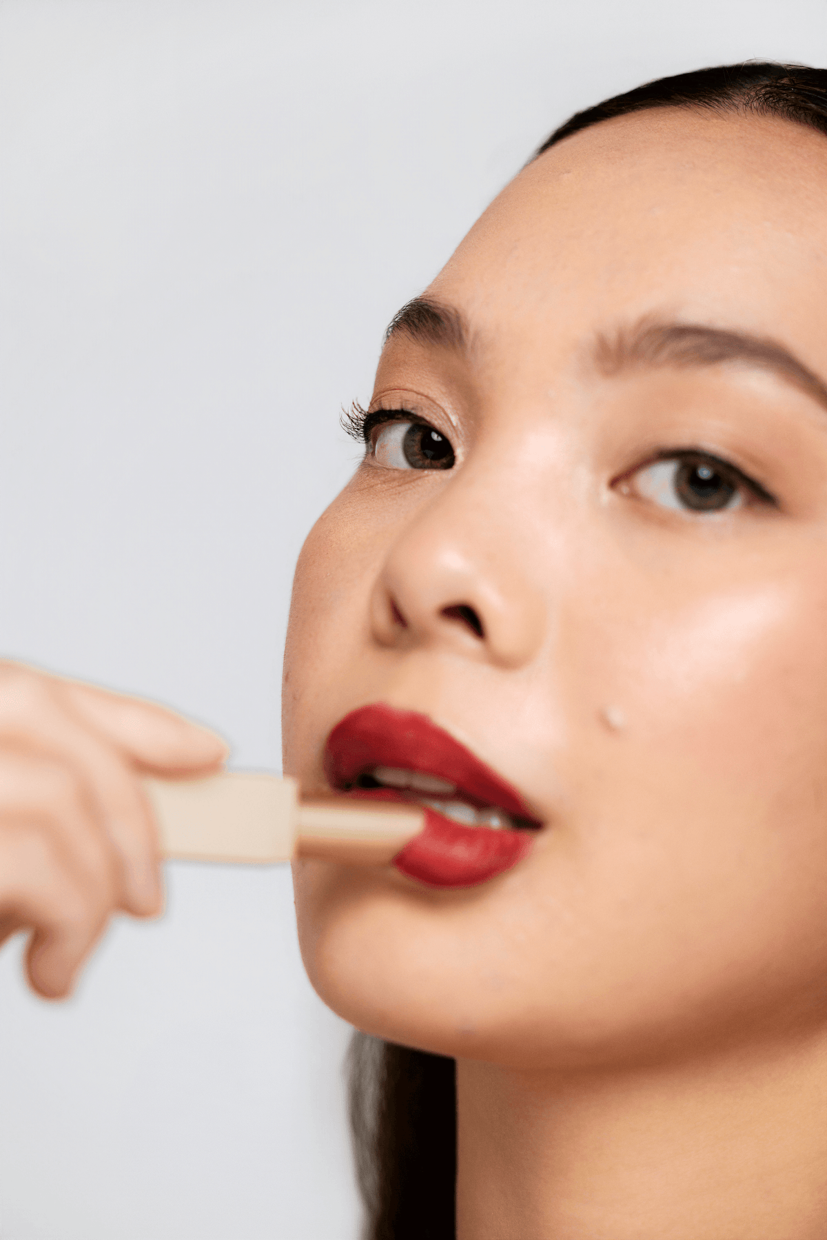 Close-up of a woman applying red lipstick with a beige applicator, showing her face and hand against a light gray background.