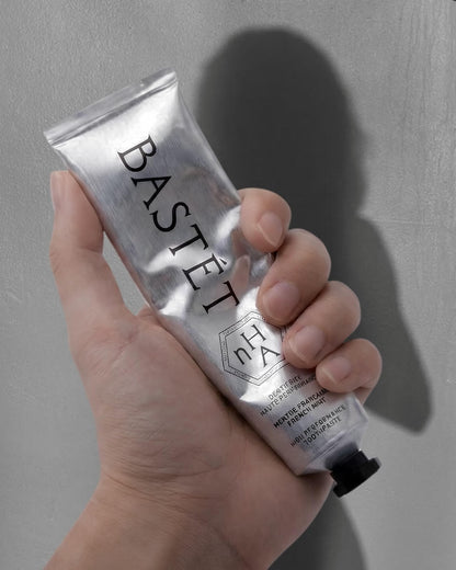 A hand holding a metallic silver squeeze tube of Bastét hand cream against a textured gray background with a soft shadow.