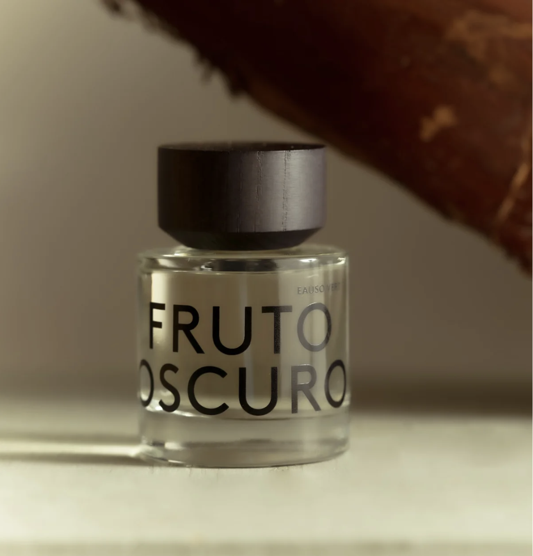 A clear glass bottle of Eauscoff 'Fruto Oscuro' with a matte black cap, set against a warm, softly lit background with a blurred brown element in the upper right.