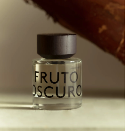 A clear glass bottle of Eauscoff 'Fruto Oscuro' with a matte black cap, set against a warm, softly lit background with a blurred brown element in the upper right.