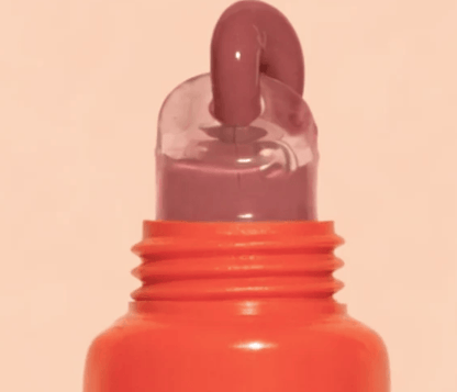 A close-up of a cosmetic tube or bottle with a bright orange-red threaded neck and a translucent cap, showing a thick glossy mauve-pink product being dispensed from the top against a pale peach background.