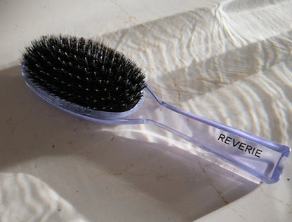 Hairbrush with black bristles and a purple handle labeled 'REVERIE' on a textured white surface.