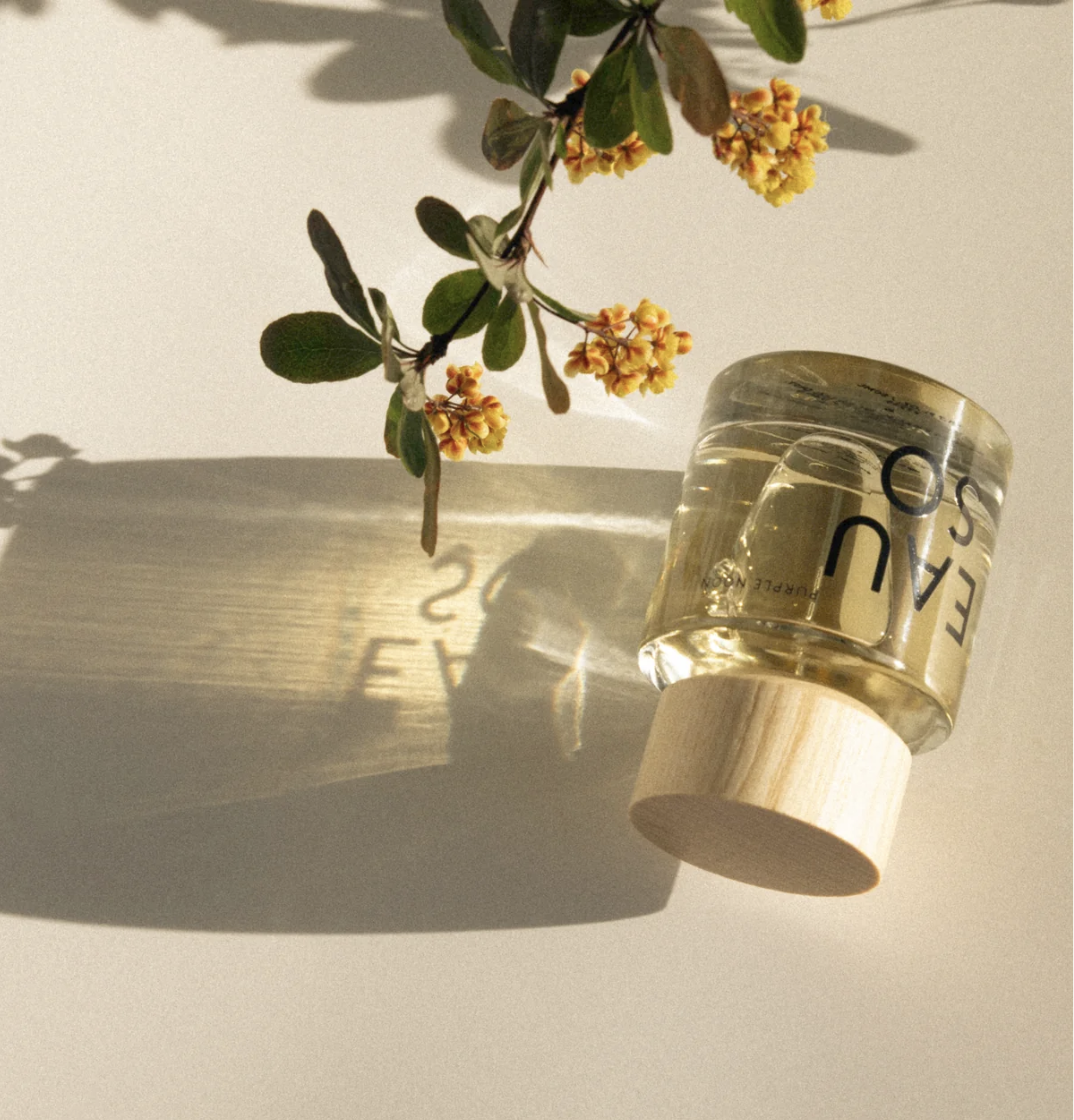 A clear perfume jar filled with pale yellow liquid, featuring black text and a light wood cap, styled on a warm beige surface with a leafy branch casting a shadow.