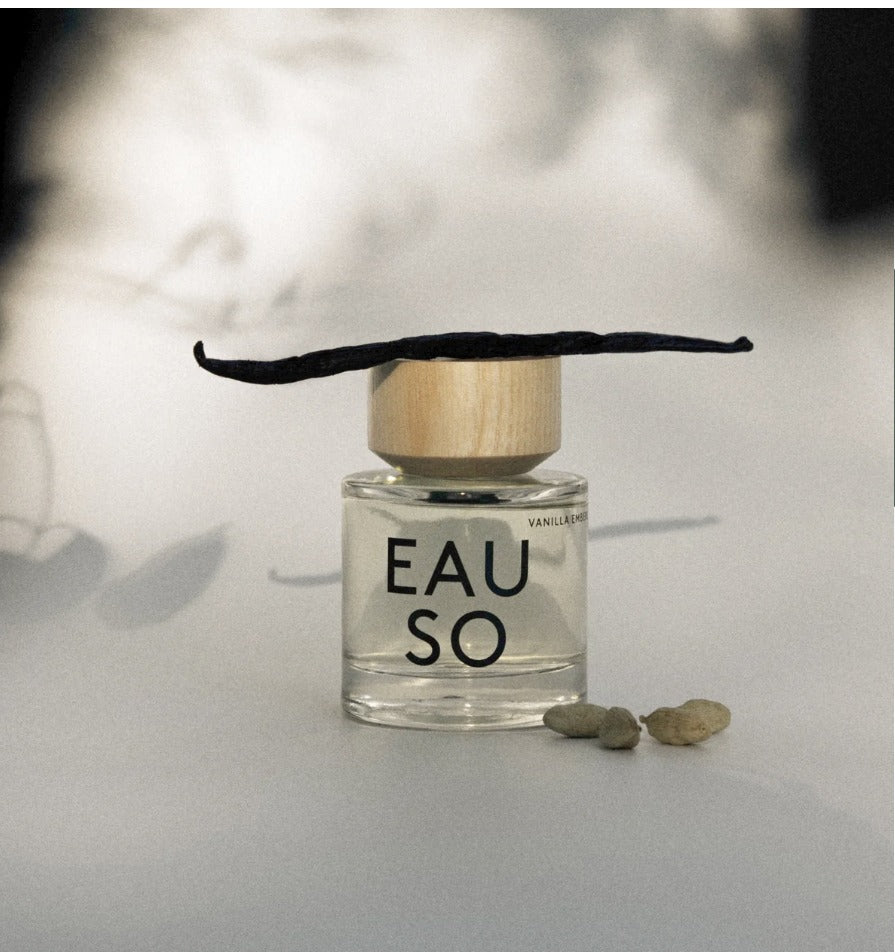 A clear glass perfume bottle labeled 'EAU SO' with a light wood cap and a dark vanilla bean resting on top, set on a light surface with a blurred warm-toned background.