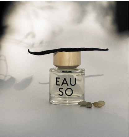 A clear glass perfume bottle labeled 'EAU SO' with a light wood cap and a dark vanilla bean resting on top, set on a light surface with a blurred warm-toned background.