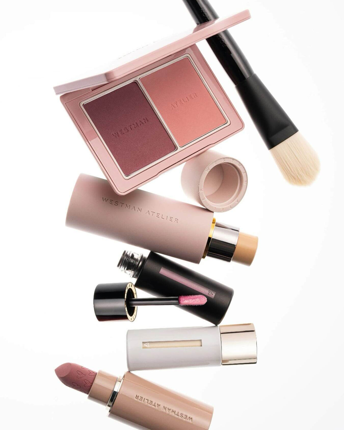 A flat-lay arrangement of Westman Atelier makeup products on a white background, including a dual-pan eyeshadow palette, a brush, a cream tube, a black-and-beige lip product, a white-and-gold tube, and a lipstick.